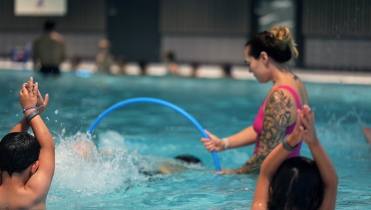 En simlärare står i en pool och håller i en rockring som barn simmar genom. 