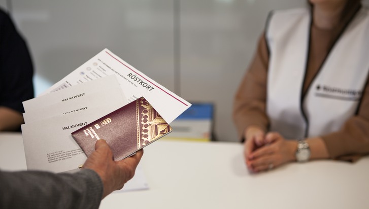 Hand med pass, röstkort och tre röstkuvert mittemot en röstmottagare i vit väst.
