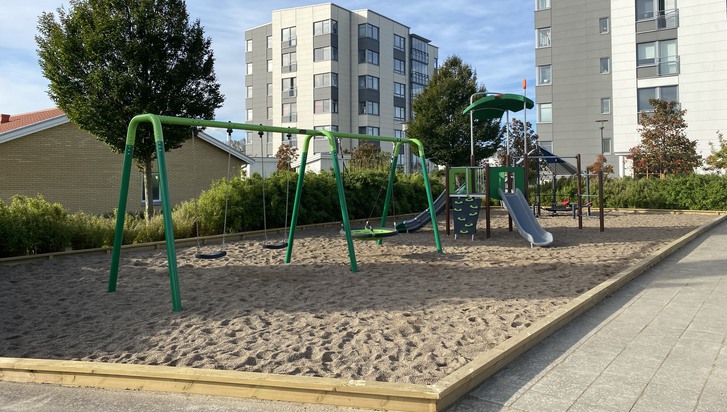 Bild som visar Sandhagens lekplats i Arlöv med sandlåda och grön gungställning med vita höghus i bakgrunden