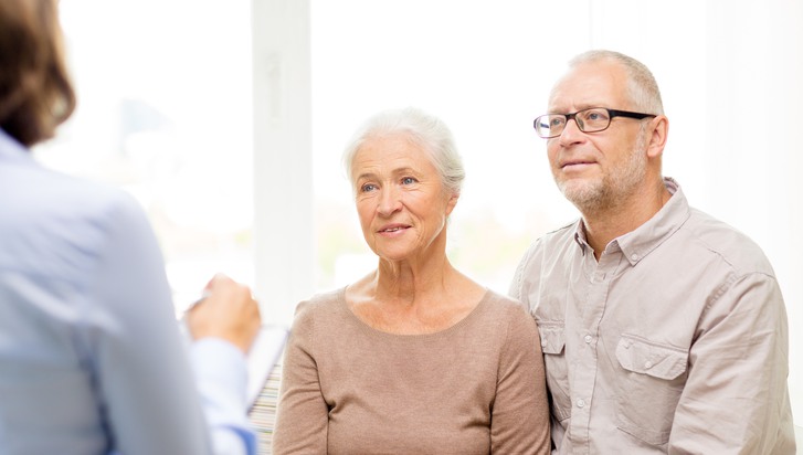 Ett äldre par, en man och en kvinna, sitter och lyssnar på när en tjänsteperson, en kvinna, pratar.