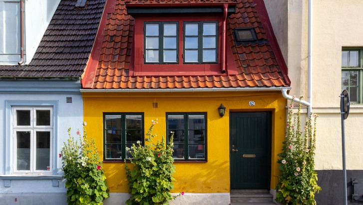 Litet gult hus med rött tak inklämt mellan två andra och större hus.