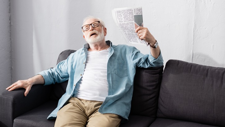 Äldre man sitter i soffa och fläktar sig med en papperstidning.