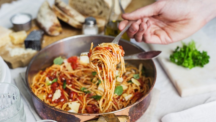 hand håller gaffel med hoprullad spaghetti ovanför gryta med spaghetti och pastasås