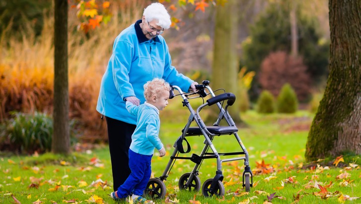 Äldre kvinna med rollator promenerar med litet barn i park. 