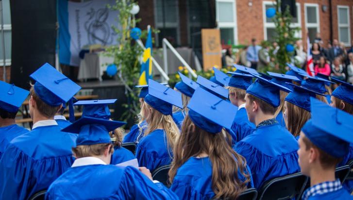 Studenter i blåa kläder och hattar sitter och tittar bort från kameran. I bakgrunden bland annat en svensk flagga.
