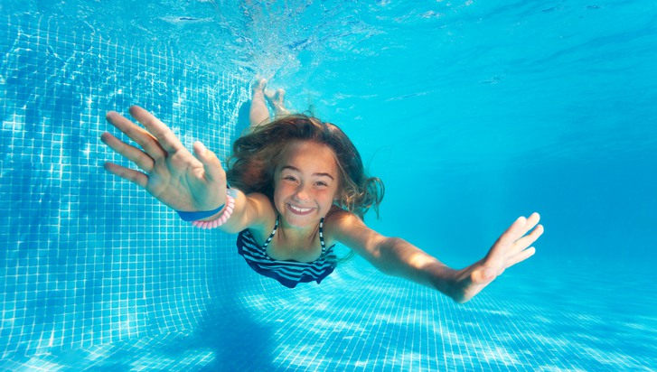 En flicka simmar under vattnet i en pool och ler mot kameran.