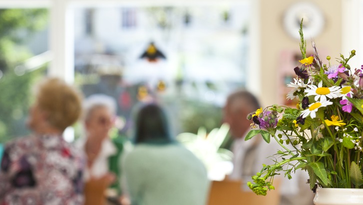 Blommor i en vas framför en grupp äldre människor som sitter vid ett bord.