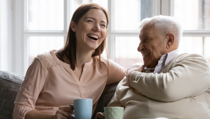 En kvinna i 30-40-årsåldern och en äldre man dricker kaffe och skrattar.